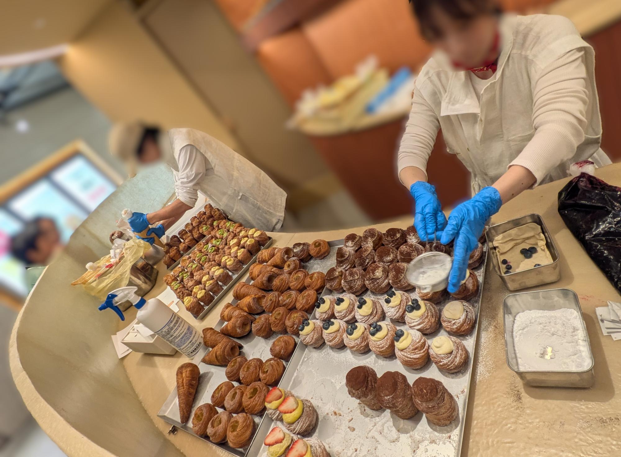 『Phi』の厨房でスイーツを仕上げる様子。粉糖をふりかける職人と、フルーツやクリームを飾ったスイーツが並ぶ作業台。