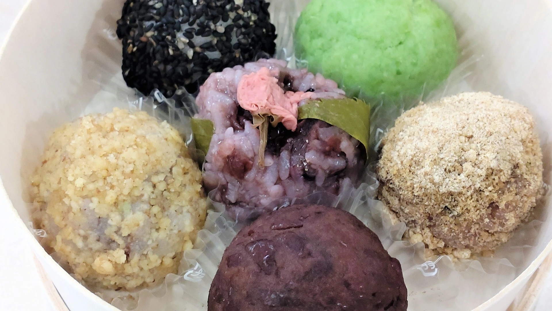 彩り豊かな6種類のおはぎが、丸い容器にバランスよく並ぶ可愛らしい一皿。黒ごま・よもぎ・きなこ・クルミ・さくら・あんこが揃う。