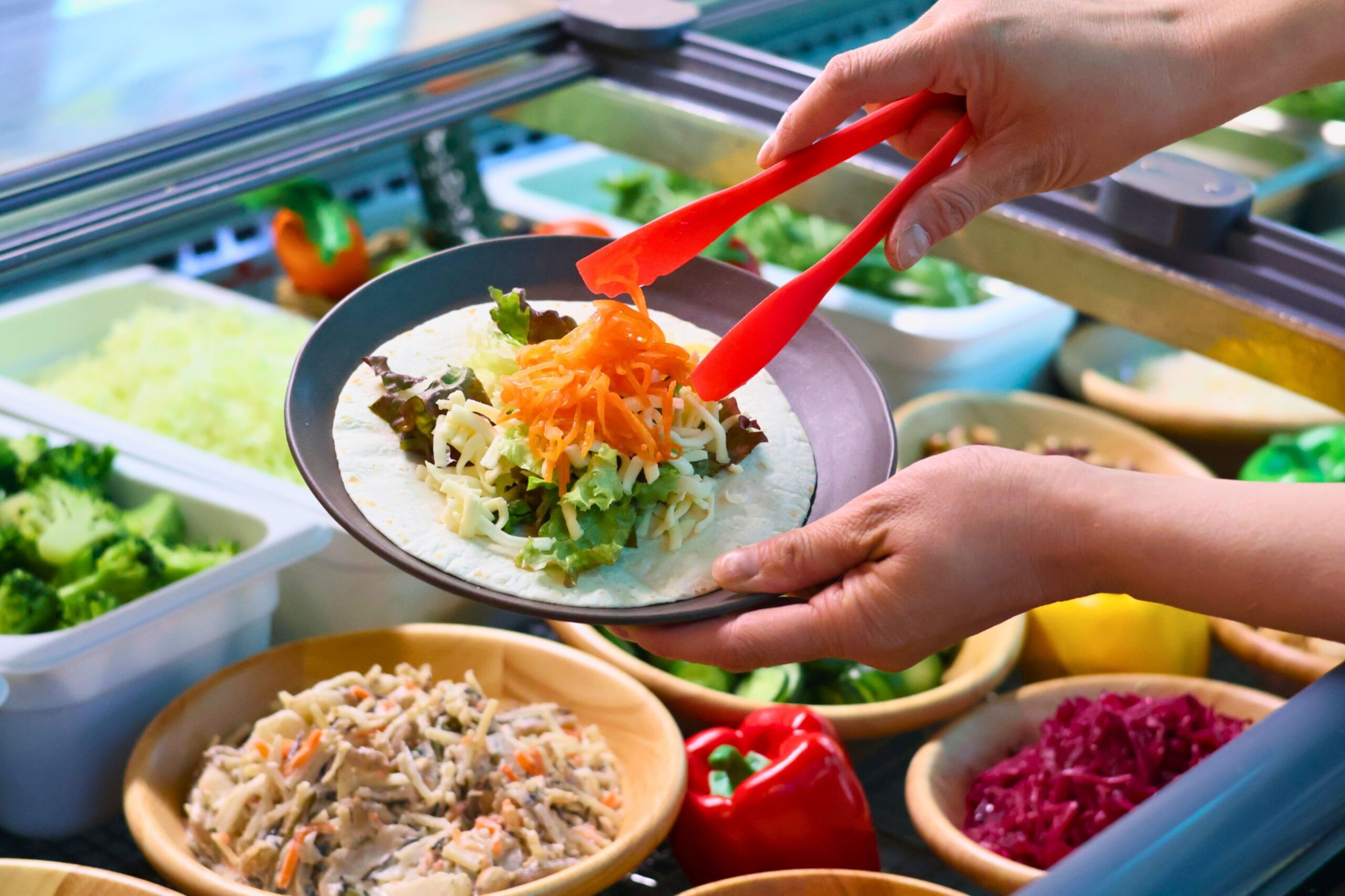 サラダバーに並ぶ野菜をトルティーヤに盛り付けている公式イメージ。