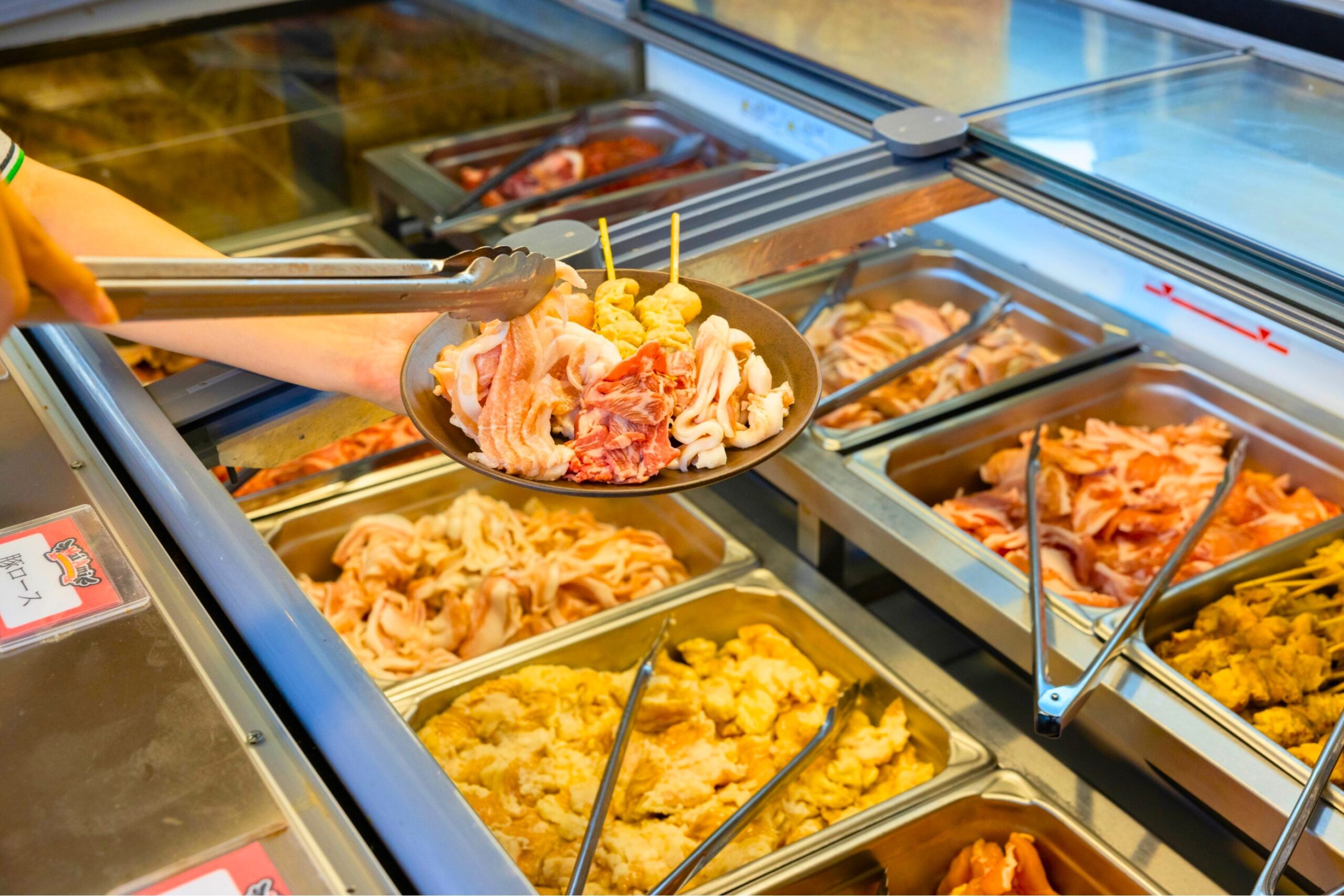 ビアガーデンのビュッフェ台に並ぶ肉類の公式イメージ。豚肉や鶏肉、串などをトングで取っている様子。