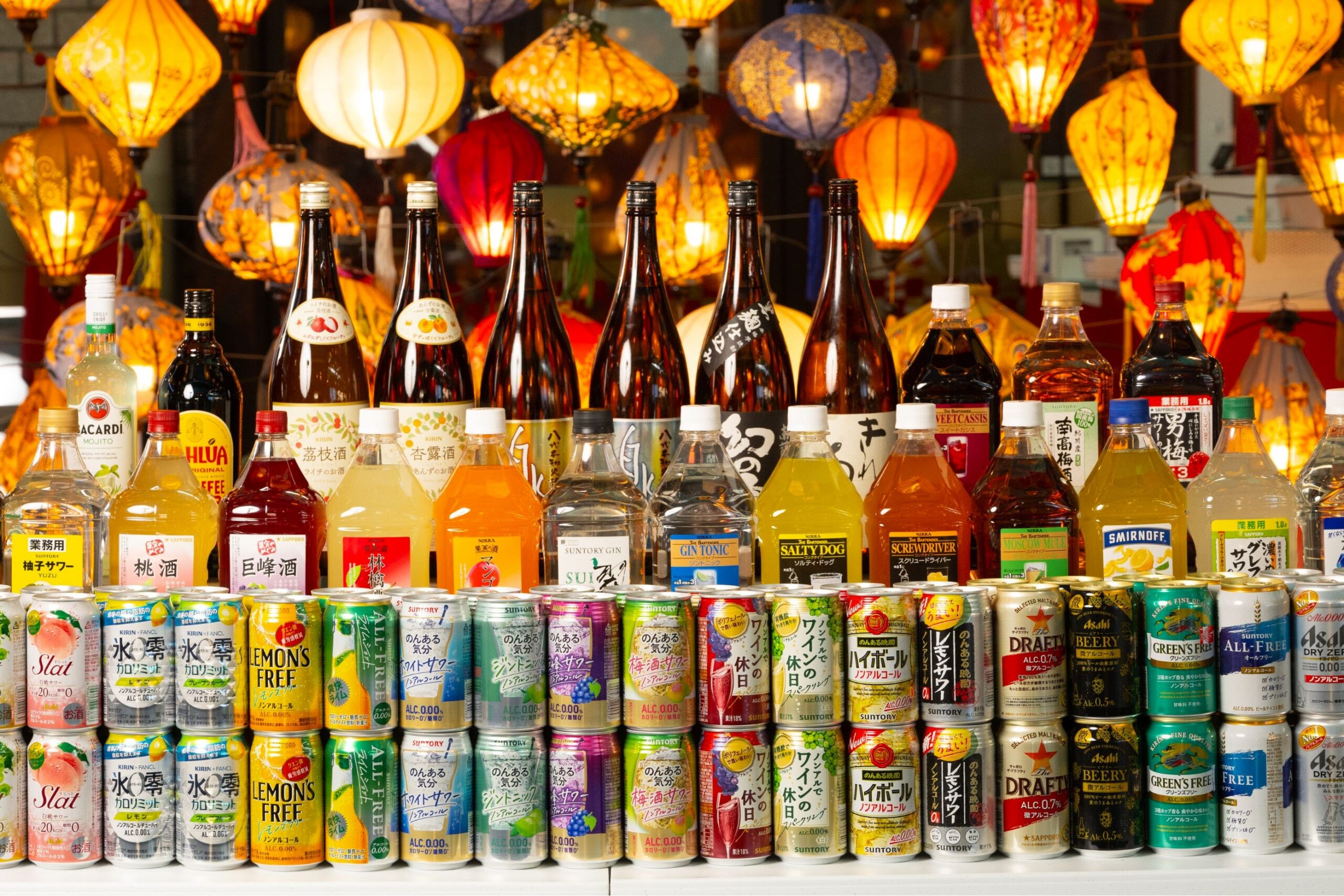 ビアガーデンのドリンクカウンターに並ぶボトルや缶ドリンクの公式イメージ。提灯の明かりが背景に映る。